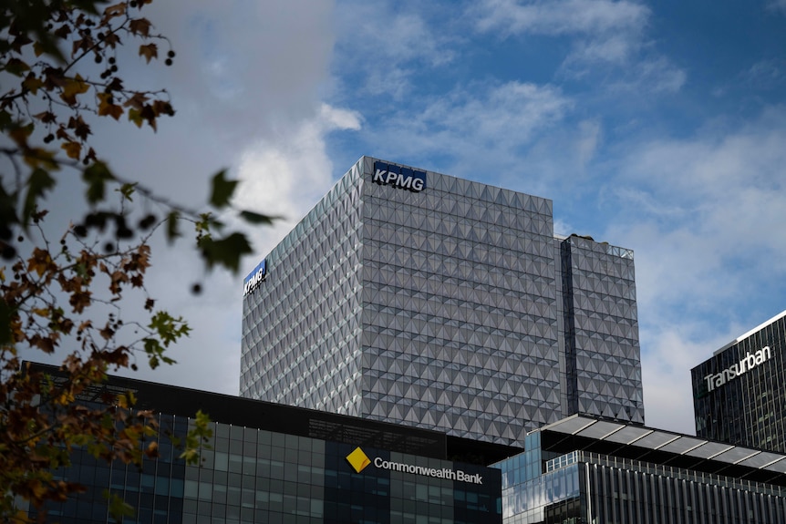 A large, grey building with a blue and white sign at the top reading KPMG. A lower Commonwealth Bank building is in front of it.