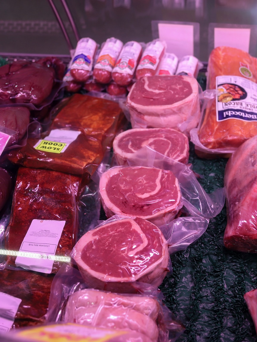 A display of a range of cuts of meat under glass and pink light in a butchers shop.
