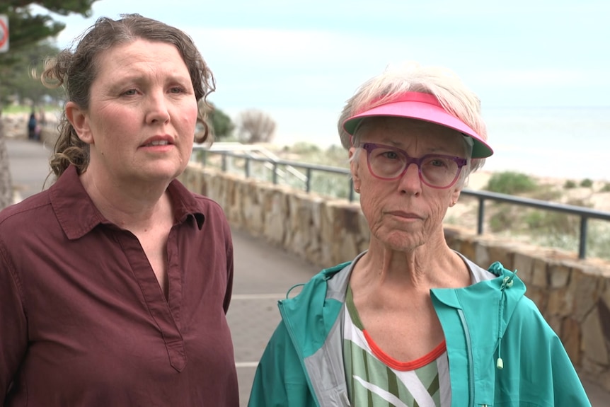 Two women look upset at the camera while standing on a footpath next to sand dunes and a beach