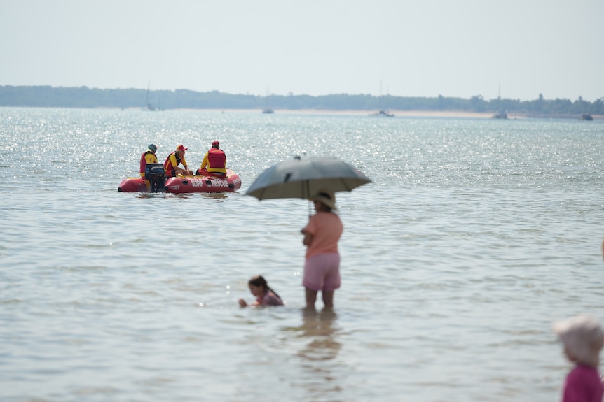 Surf life savers on a rubber ducky in the water with a person standing in the foreground holding an umbrella in the water