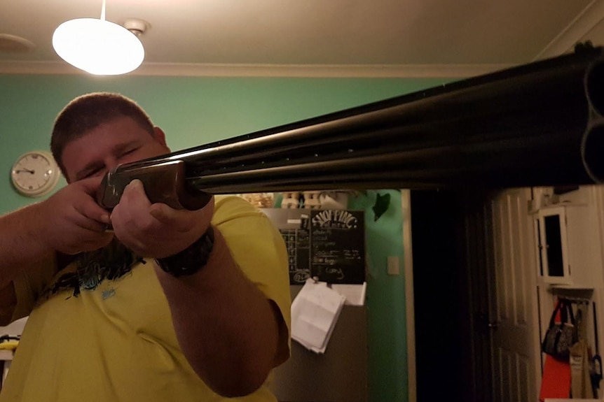 A man with cropped, dark hair aims a longarm weapon while standing in a kitchen.