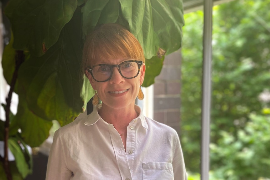A middle-aged woman with strawberry blonde hair and black-rimmed glasses in a white shirt smiles at camera in a room