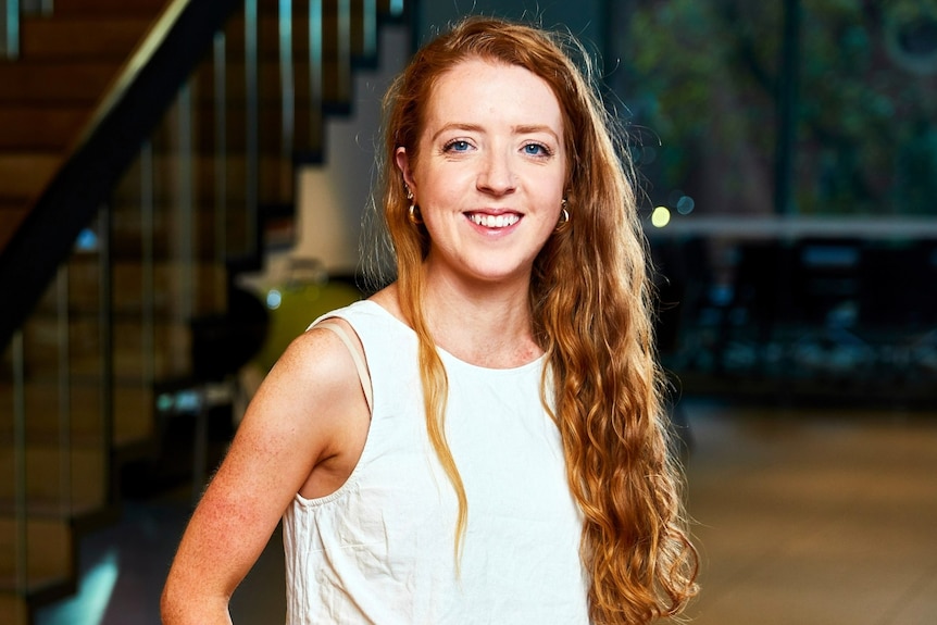 A headshot of a young woman in a white linen sleeveless shirt, smiling. She has long red hair.