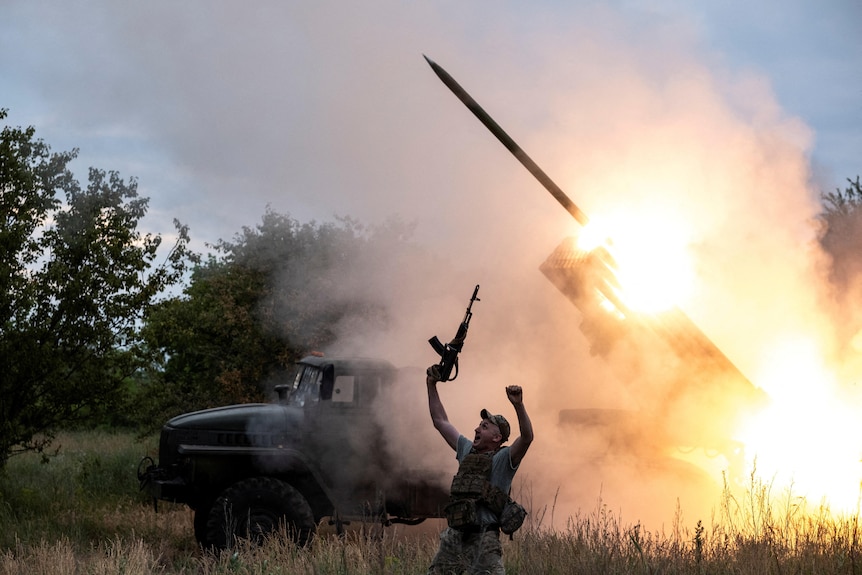 A soldier raises his hands, one of them holding a machine gun, as behind him a missile launches from the back of a truck.