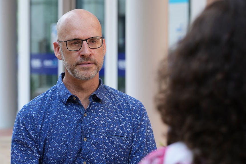 A man in a button-up shirt and glasses wearing a serious facial expression as he speaks to a journalist.