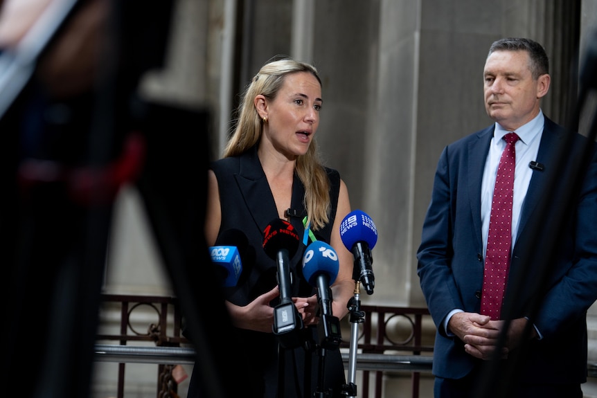 A woman speaks to microphones while a man looks on