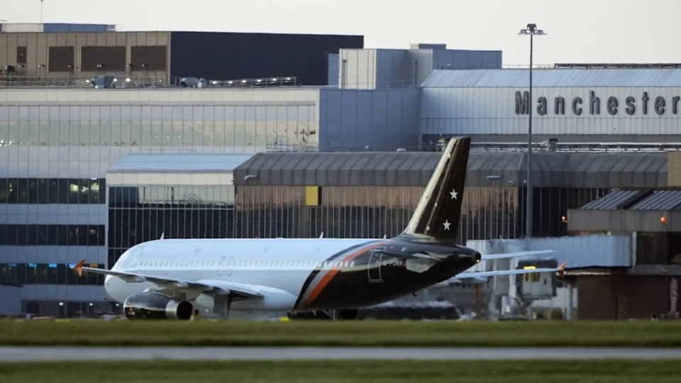 An airplane is on the runway, with grass in the foreground and an airport in the background with Manchester emblazoned on it