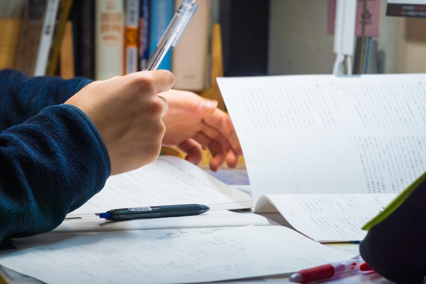 A close up image of white hands hovering over a notebook and a textbook, with a pen in hand.