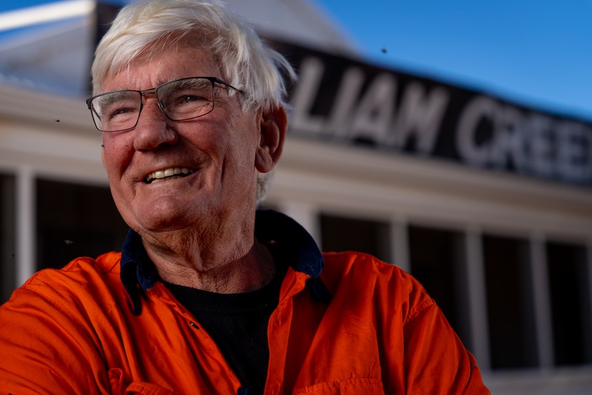 A close-up of a man smiling at the camera, infront of a sign reading "william creek hotel"