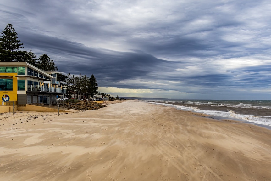 Dark grey clouds above a sweeping sandy beach and waves along the coast of Henley Beach. Waves crash against the sand.