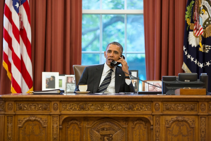Obama sits behind the desk and holds a black phone receiver to his ear