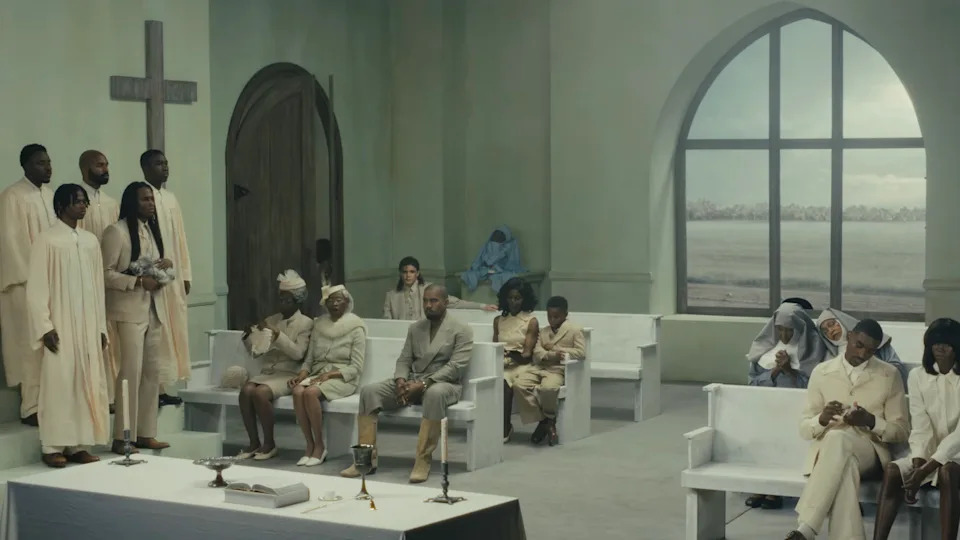A group of people in a minimalist church setting, wearing white and beige clothing, with a large window and cross in the background. A still from Kanye West music video "Father."