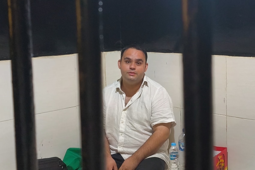a man in a white dress shirt and black pants sits in a jail cell photo taken through bars