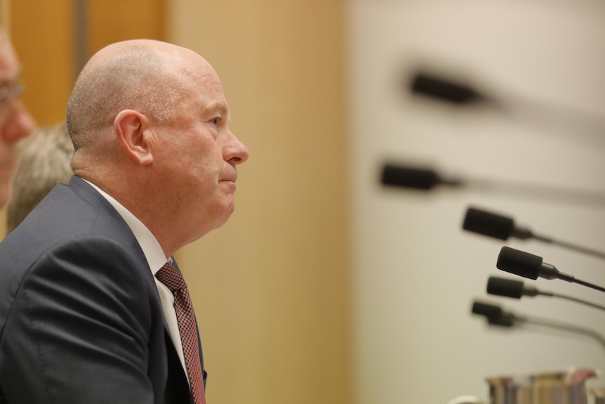 A bald man in a suit and red tie speaks into a microphone while siting at a desk in a room decorated with timber.