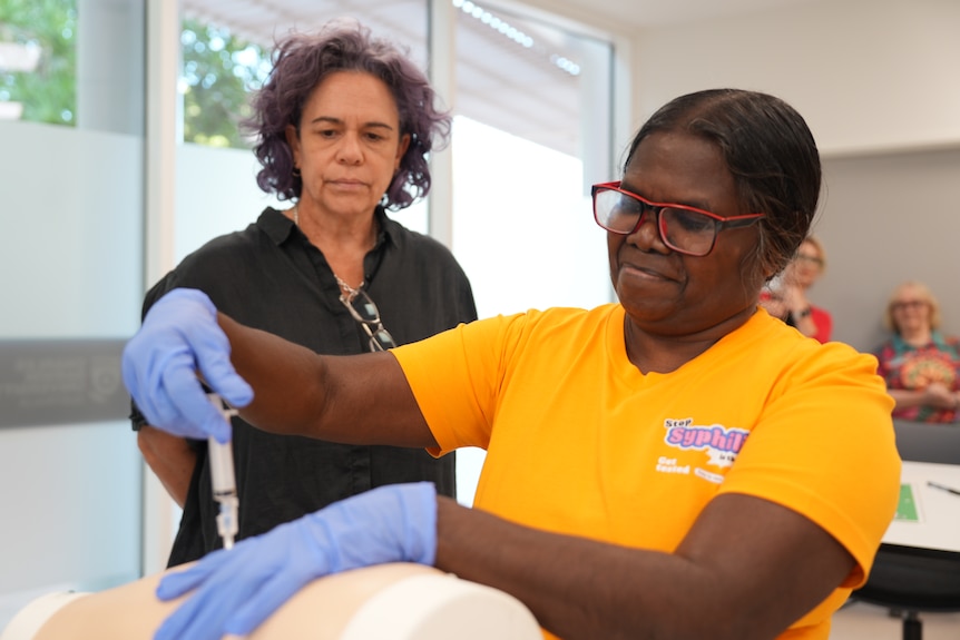 An Indigenous woman injecting a test dummy with a needle, she is wearing glasses and rubber gloves.