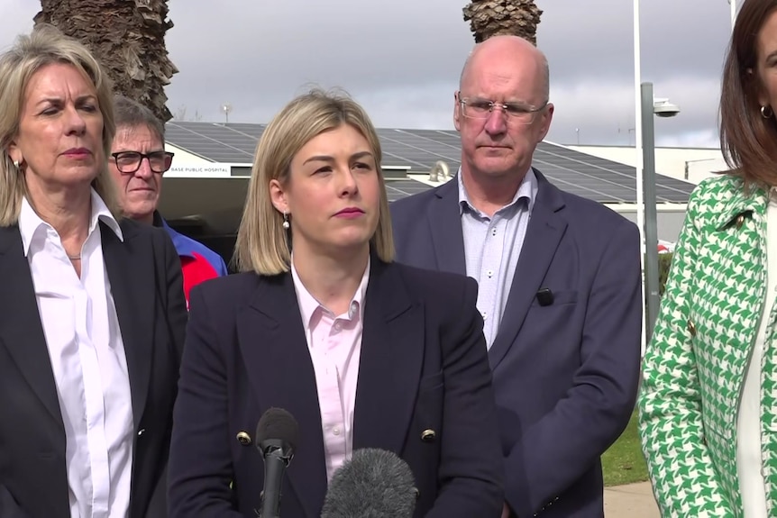 A blonde woman wearing a black suit jacket and pink shirt speaks in front of a microphone.