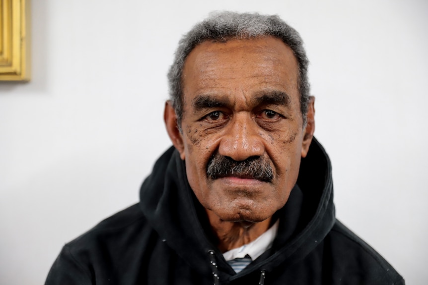 Man with moustache wearing black hoodie stares directly into camera in front of white wall