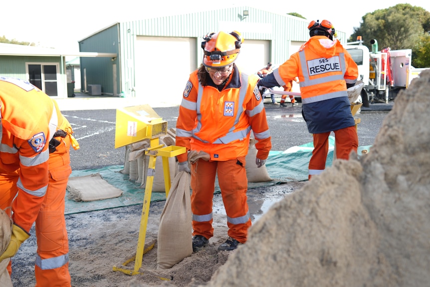A man wearing orange filling a sandbag with a yellow machine in front of sheds