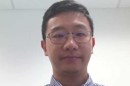 A head and shoulders shot of an Asian-Australian man, with black hair, wearing spectacles and a blue and white business shirt.