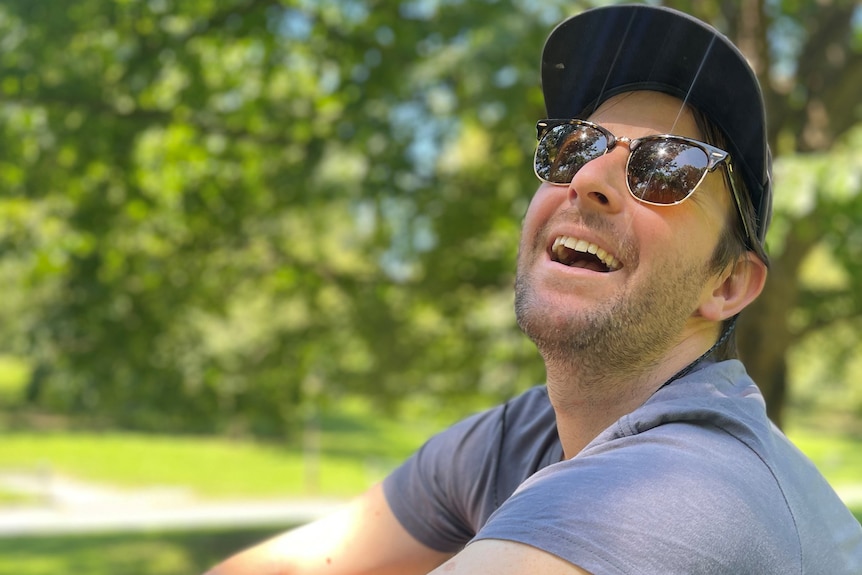 Laurence wears sunglasses and hat in a park as he laughs