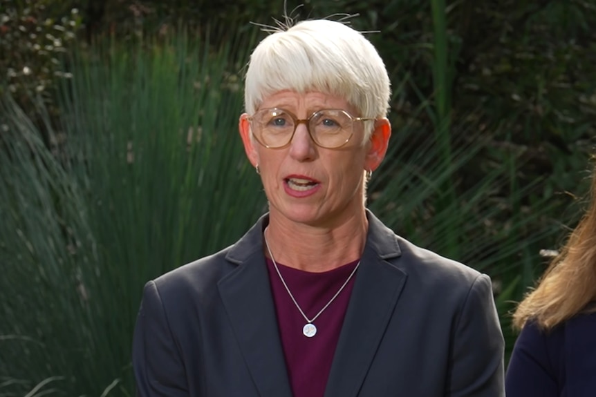 Ms Coombe with short white hair, glasses, a grey blazer, speaking in front of a bush.
