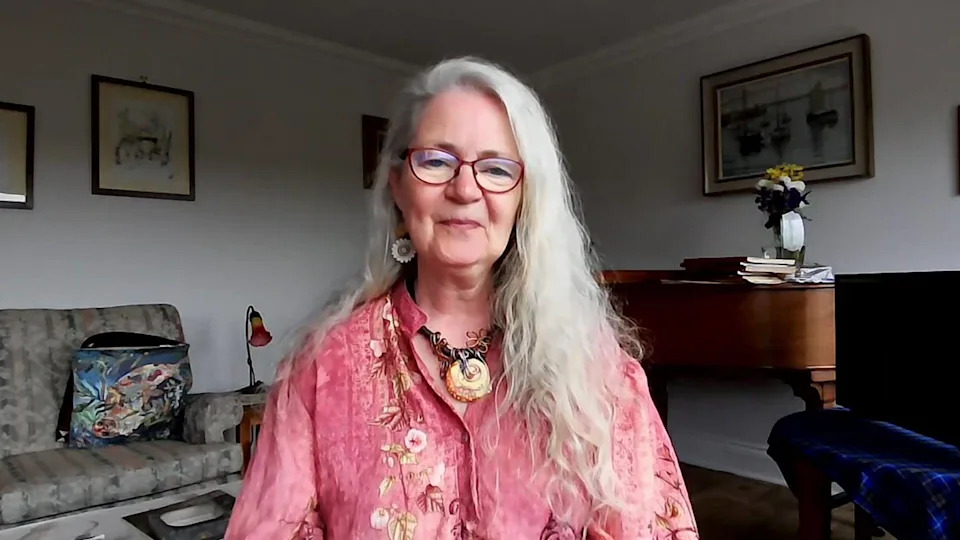 A woman with long white hair and glasses sits facing the camera in a softly lit living room, wearing a pink patterned shirt and a large pendant necklace. Behind her, a wooden desk holds stacked books and a vase of flowers, with framed artwork on the walls and a patterned armchair visible to the left.