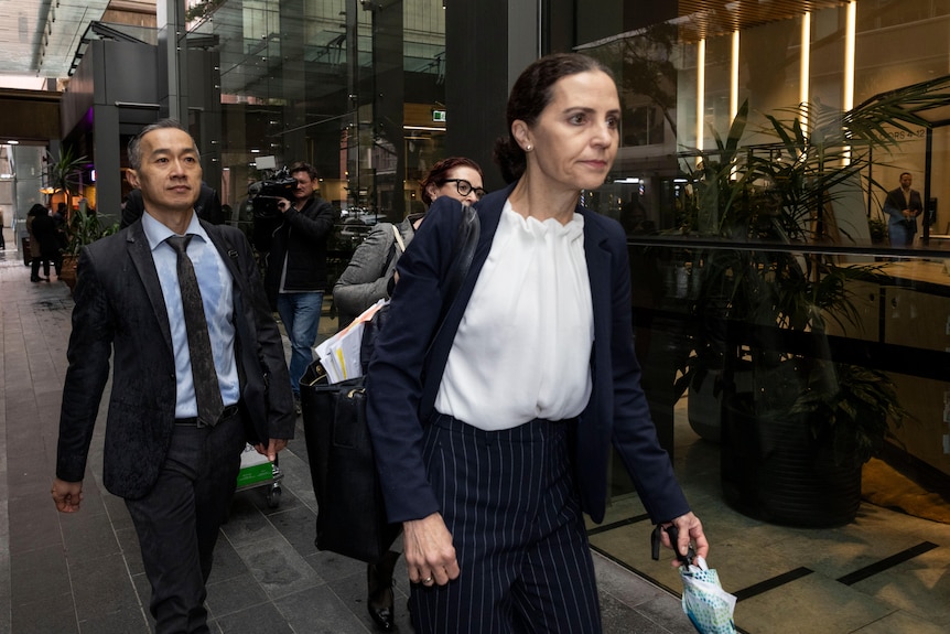 Ms Wicks in a white shirt, blazer and pants walking on a street path holding an umbrella.