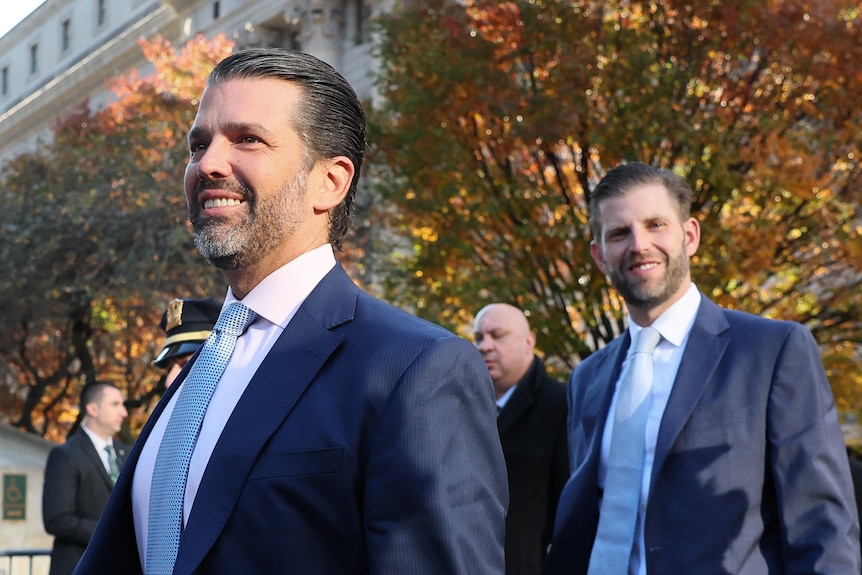 Donald Trump stands in front of Eric Trump. Both of them are smiling. There's trees in the background.