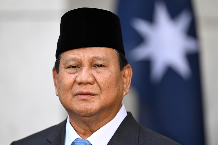 Prabowo Subianto wearing a dark suit, white shirt and black hat standing in front of a blurred Australian flag
