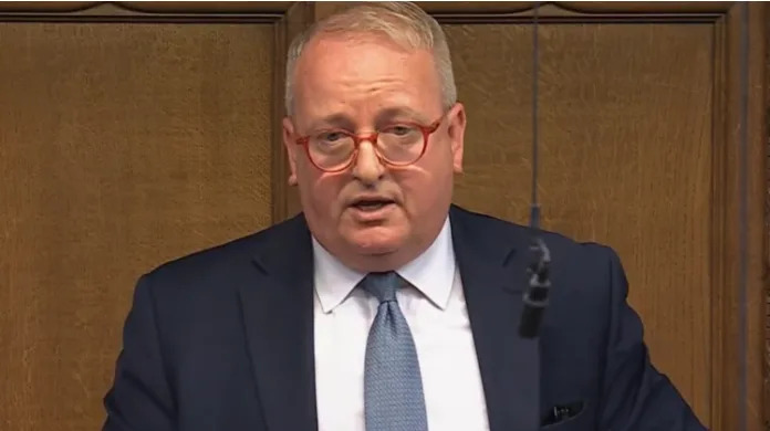 A man with short fair hair and red-framed glasses stands in the House of Commons in a navy blue suit, white shirt and blue tie. The wood panelling of the chamber is behind him, and a microphone hangs on a wire in front of him.