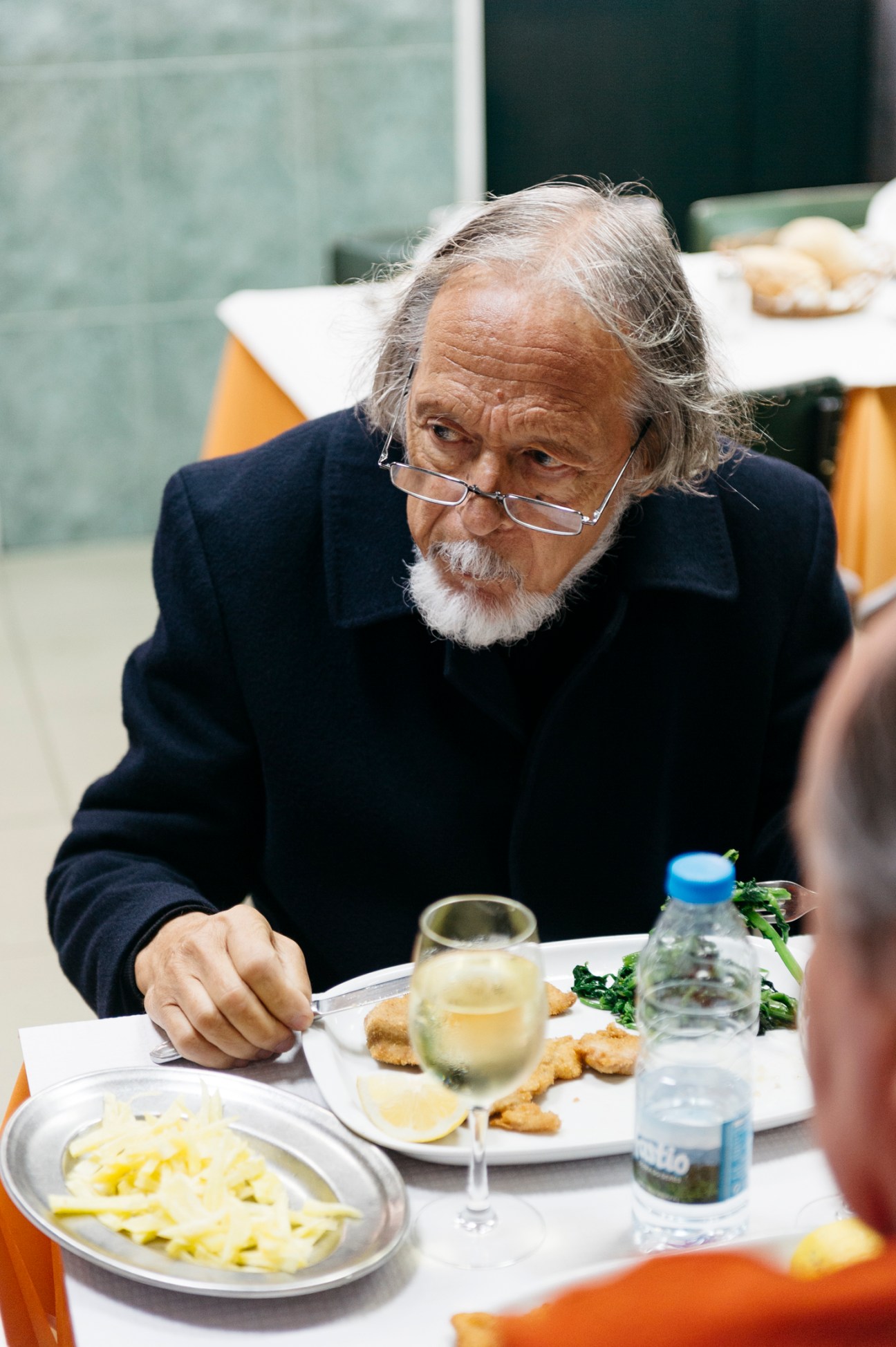 Elderly man with glasses and white beard eating at table with wine glass and plate of food