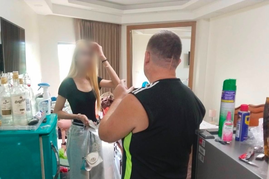 A woman talks to a man in a messy room with alcohol bottles on a cupboard.