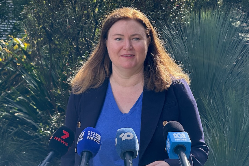 Tara Moriarty with long brunette hair, a navy blazer and bright blue top, speaking in front of a bush.
