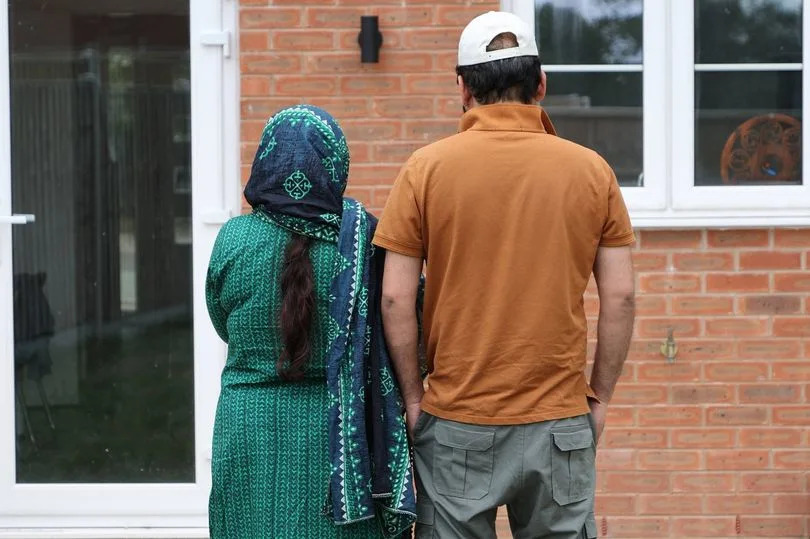 An adult male and female stand outside in front of a red brick house, with the male wearing a hat and the female wearing a headscarf.