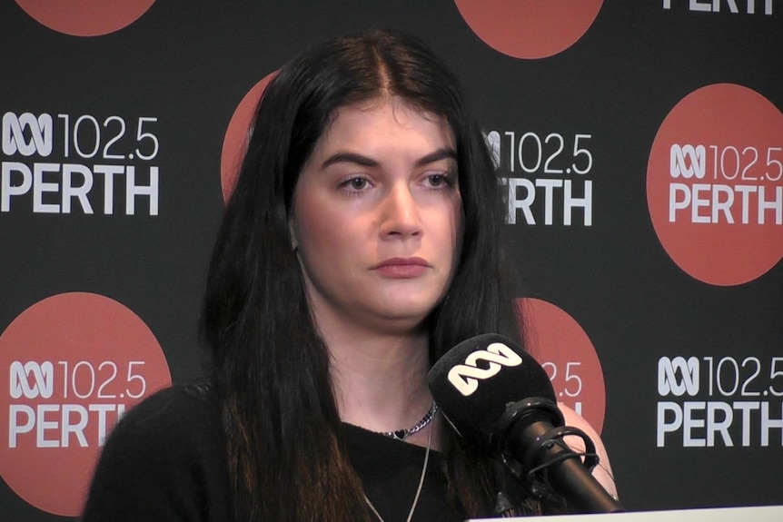 A woman in a black shirt stands in front of a microphone in a studio