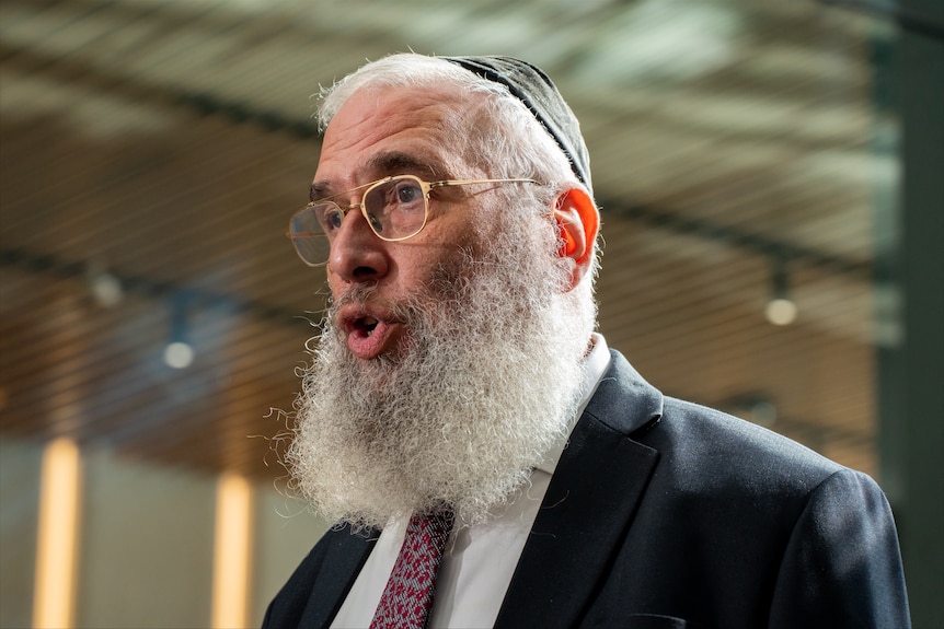 Rabbi Kastel in a black suit and red tie with a bushy beard as well as a kippa walks and talks to media.
