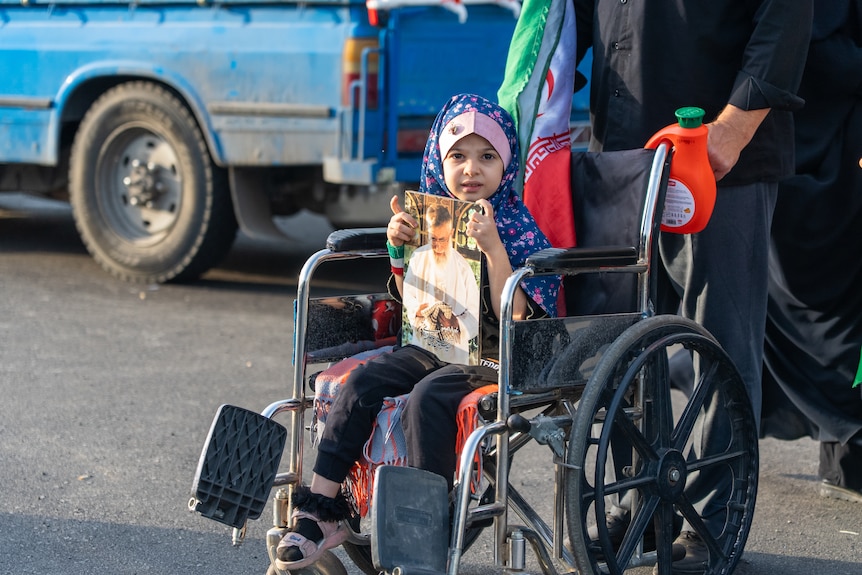 A young girl in a colourful hijab sits in a wheelchair and holds a photo of the late Ayatollah.