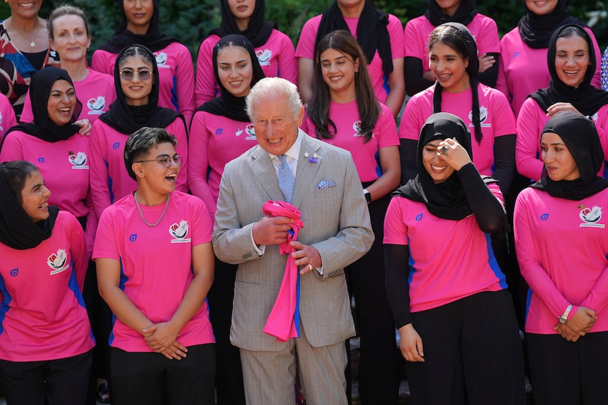 An elderly man in a grey suit smiles in a crowd of young female cricket players.