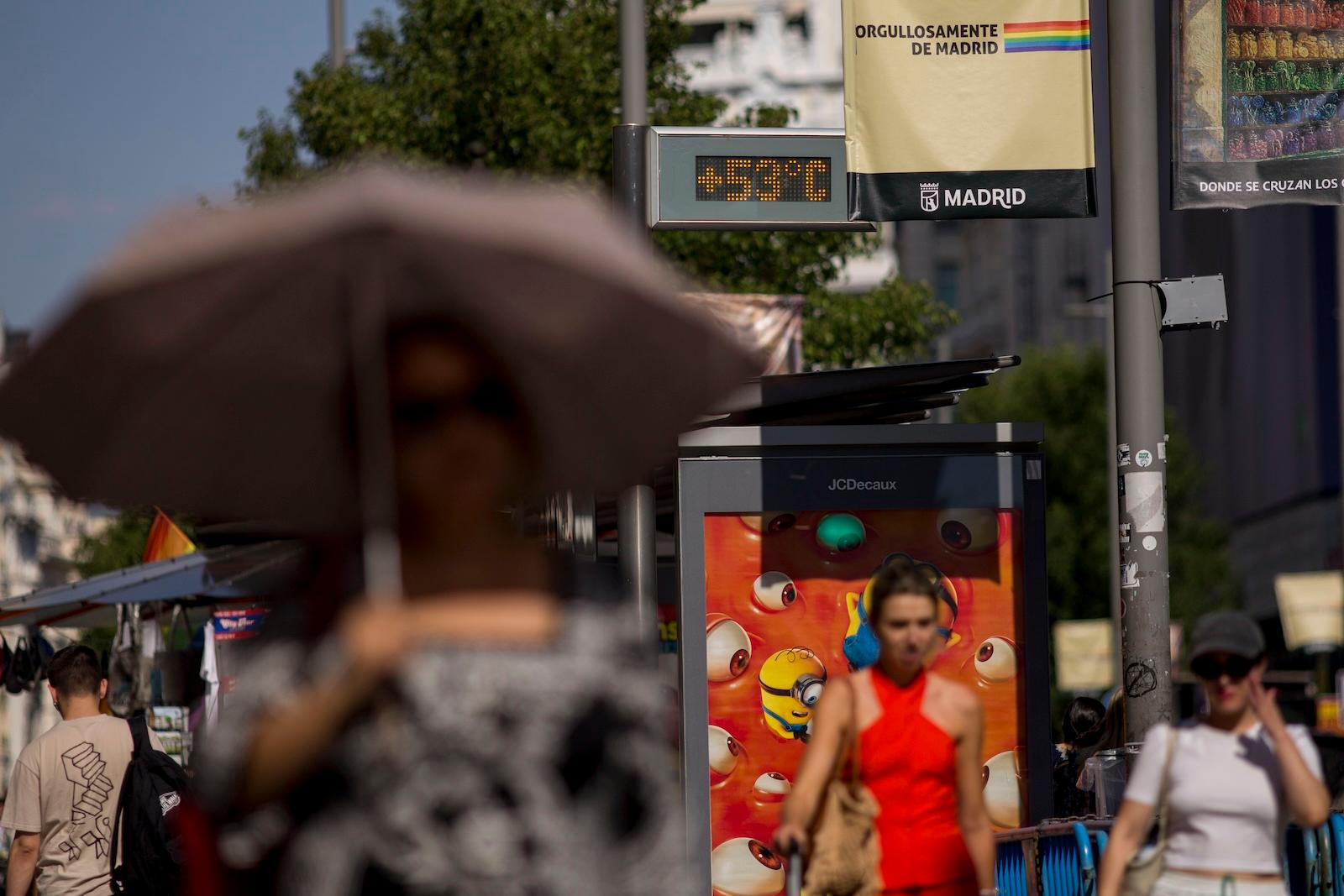 people under umbrellas walk through a seemingly busy area passing a busstop to their left with a thermometer screen that reads +53 degrees C