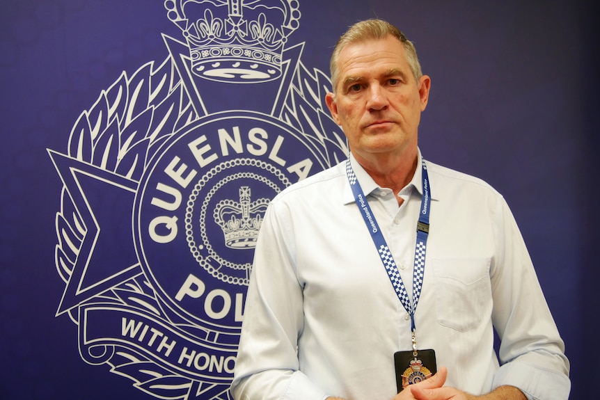 A middle-aged man in a business shirt wears a police badge on a lanyard as he stands in front of a Queensland Police backdrop.