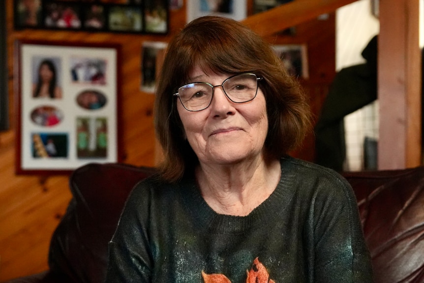 A woman smiles into the camera, sitting inside wearing glasses.