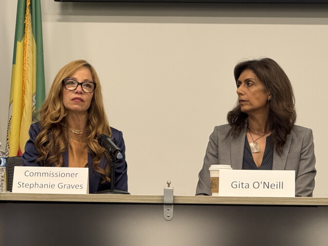 Two women sit at a desk. The one on the left has long light colored hair and is wearing glasses. Her name tag reads Stephanie Graves. The one on the right is wearing a gray suit jacket. Her name plate reads Gita O'Neill.