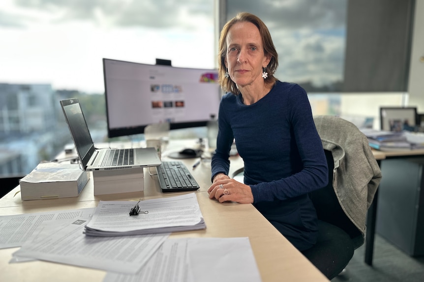 Heather Douglas sits at a desk with a laptop and computer screen with a large window behind her