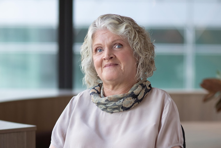 A woman with chin length white wavy hair wears a scarf over a cream top and sits in a chair in a glassed room.