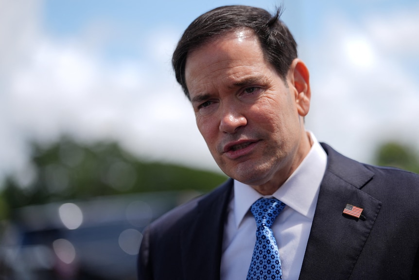 Marco Rubio speaking to reporters outside, wears blue suit, light blue tie with polka dots, US flag on lapel.