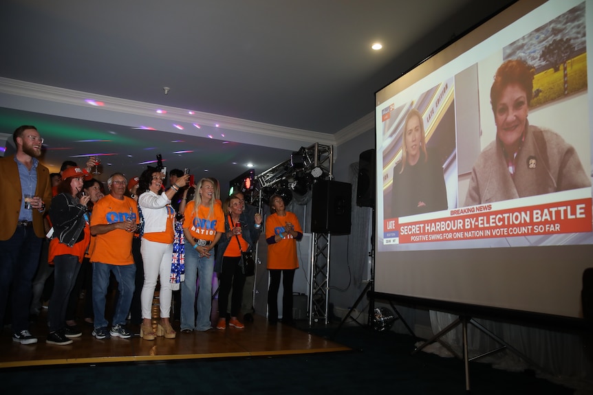 People watch a big screen with Pauline Hanson on it.