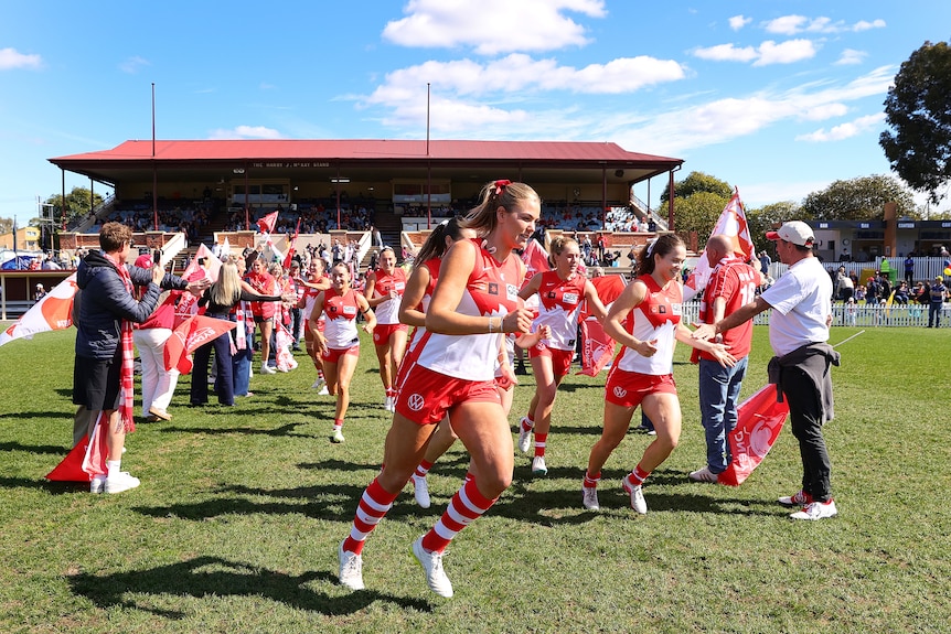 Female AFL players in red and white run out onto a green pitch on a sky blue day.