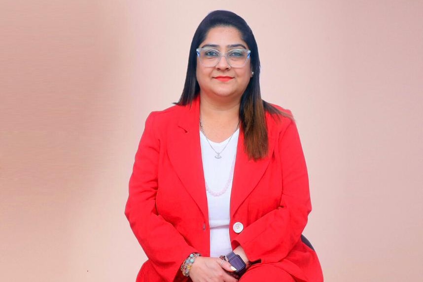 Souparna wears a red jacket and pants, white top, sits on a stool, smiles, wears glasses, professional