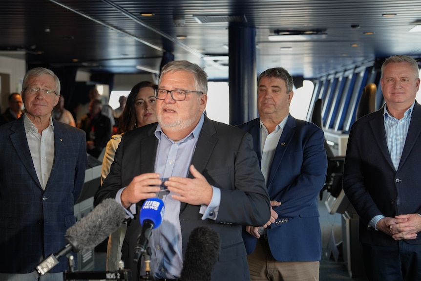 A man with short grey hair addresses media while wearing a blue suit and standing in the bridge of a ferry.