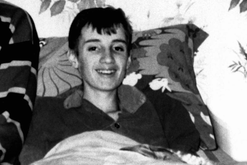 Black and white image of a teenager with short hair seated, smiling, slumped on a couch.